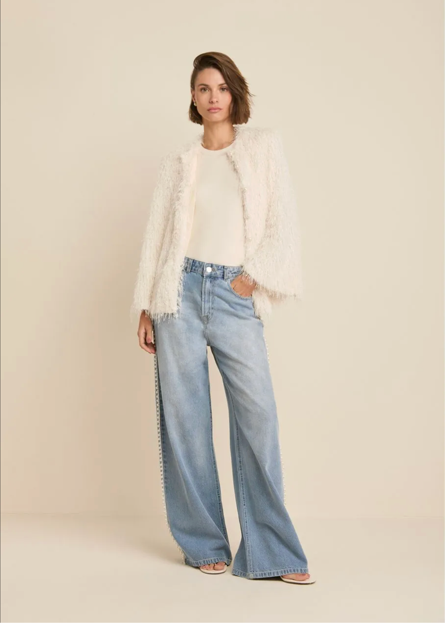A model stands against a plain beige studio background wearing a cream fuzzy cropped jacket over a cream top, paired with light-wash wide-leg jeans and flat sandals.