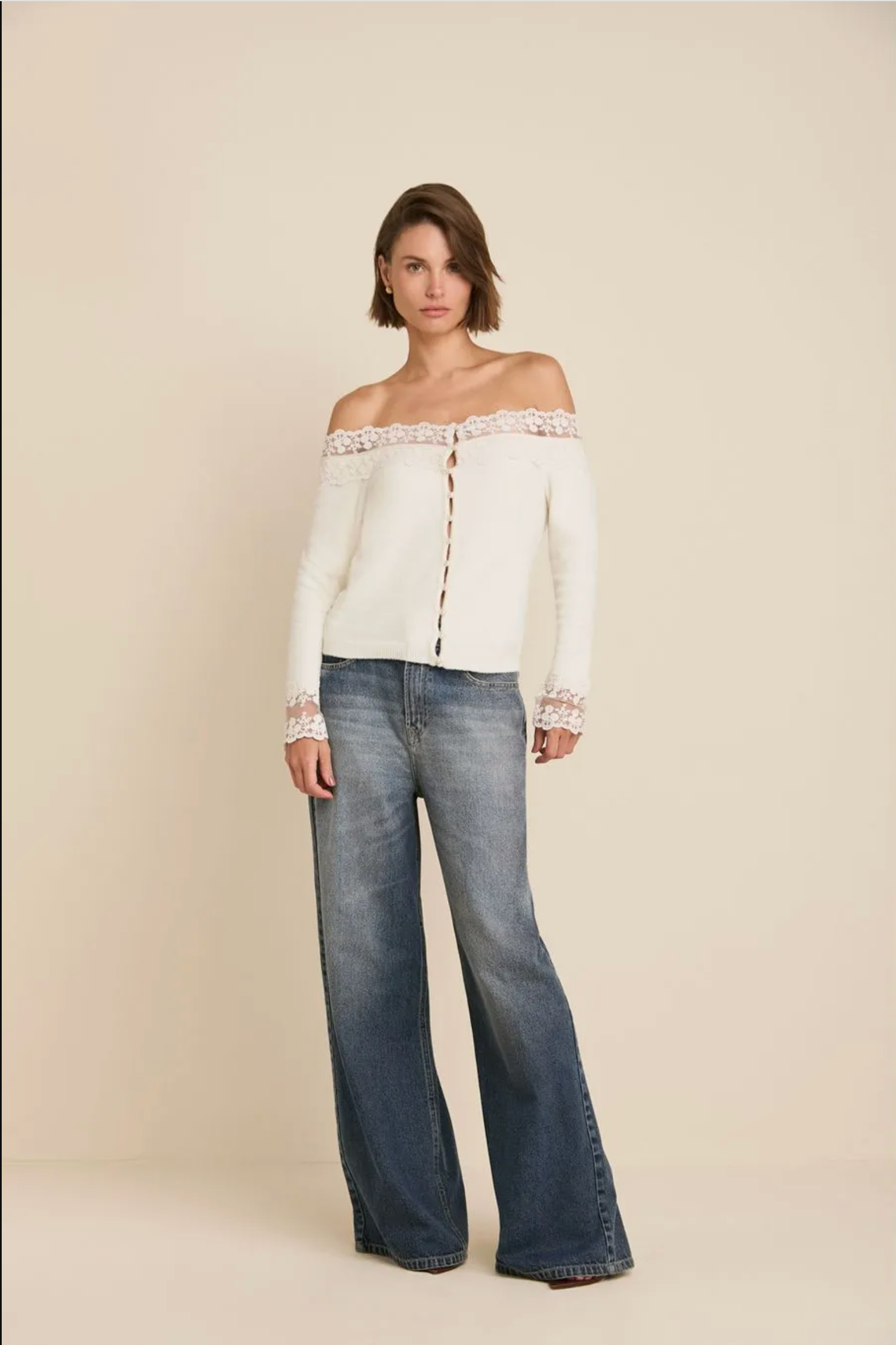 A woman modeling an off-the-shoulder cream knit top with lace trim and blue wide-leg jeans against a solid beige studio background.