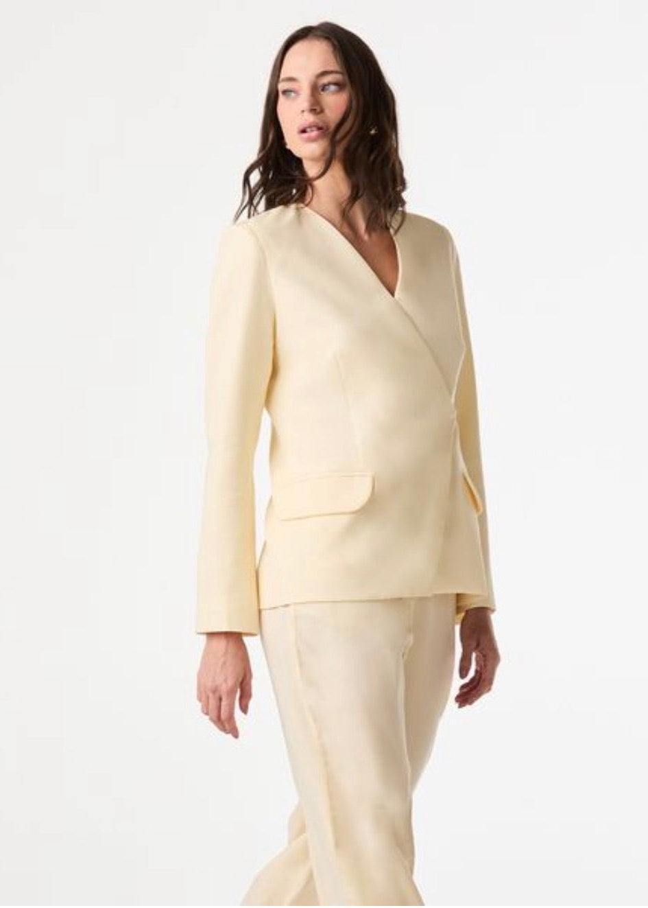 A woman modeling a light cream or pale yellow two-piece suit with a wrap-style blazer and matching trousers against a plain white studio background.