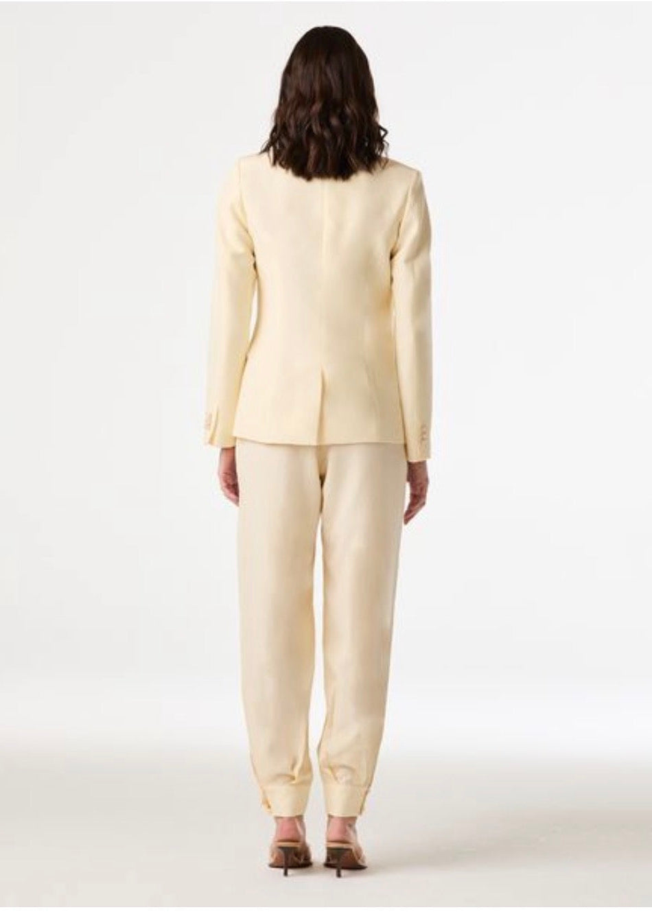 A model viewed from the back wearing a light cream or pale yellow two-piece suit with a blazer and matching trousers, paired with brown heels, against a plain white studio background.