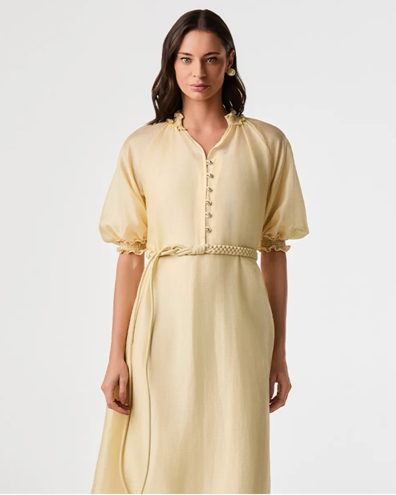 A woman modeling a pale yellow midi dress with short puff sleeves, a buttoned neckline, and a braided belt at the waist, photographed against a plain light gray studio background.