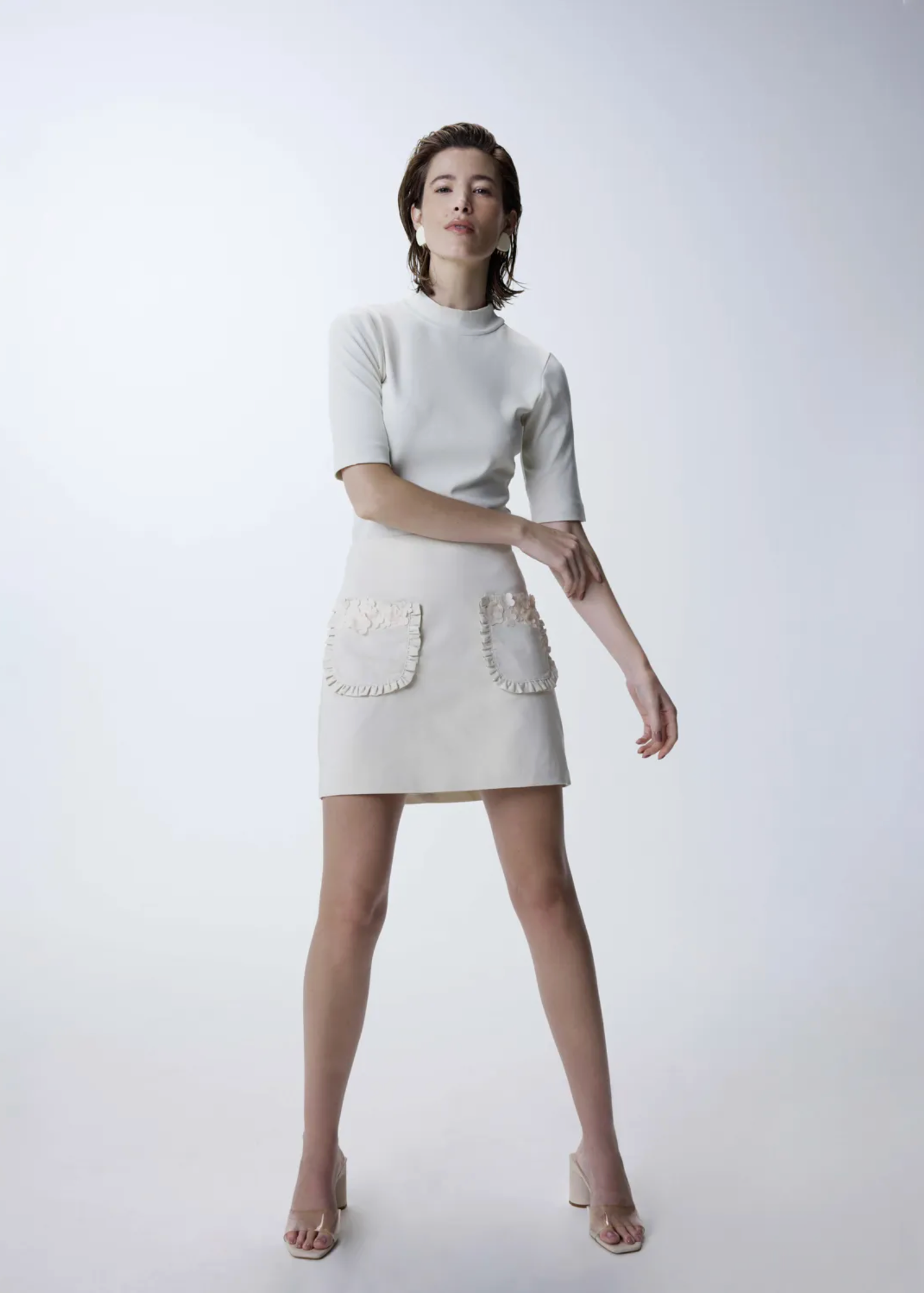 A model stands against a light yellow studio background wearing a cream-colored short-sleeve mock-neck mini dress with two decorative pearl-embellished pockets on the skirt, paired with nude high-heeled sandals.