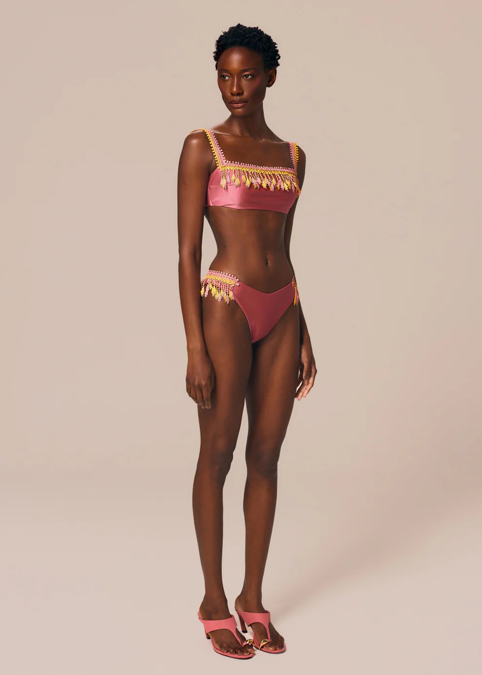 Woman wearing a pink bikini with gold fringe details on a beige background