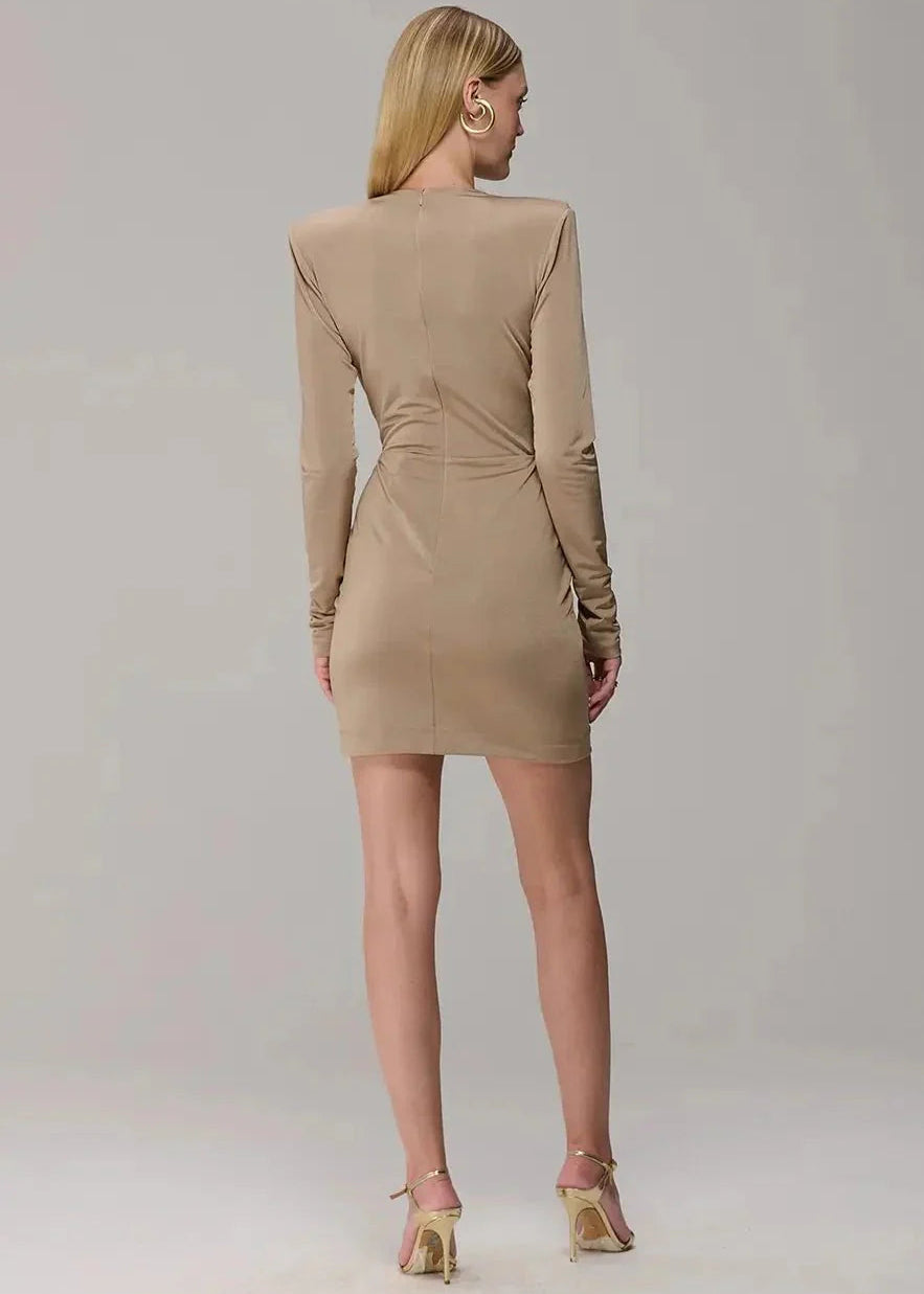 A woman viewed from the back wearing a fitted beige long-sleeve mini dress with structured shoulders and a ruched waist, paired with metallic gold high-heeled sandals against a light gray studio background.