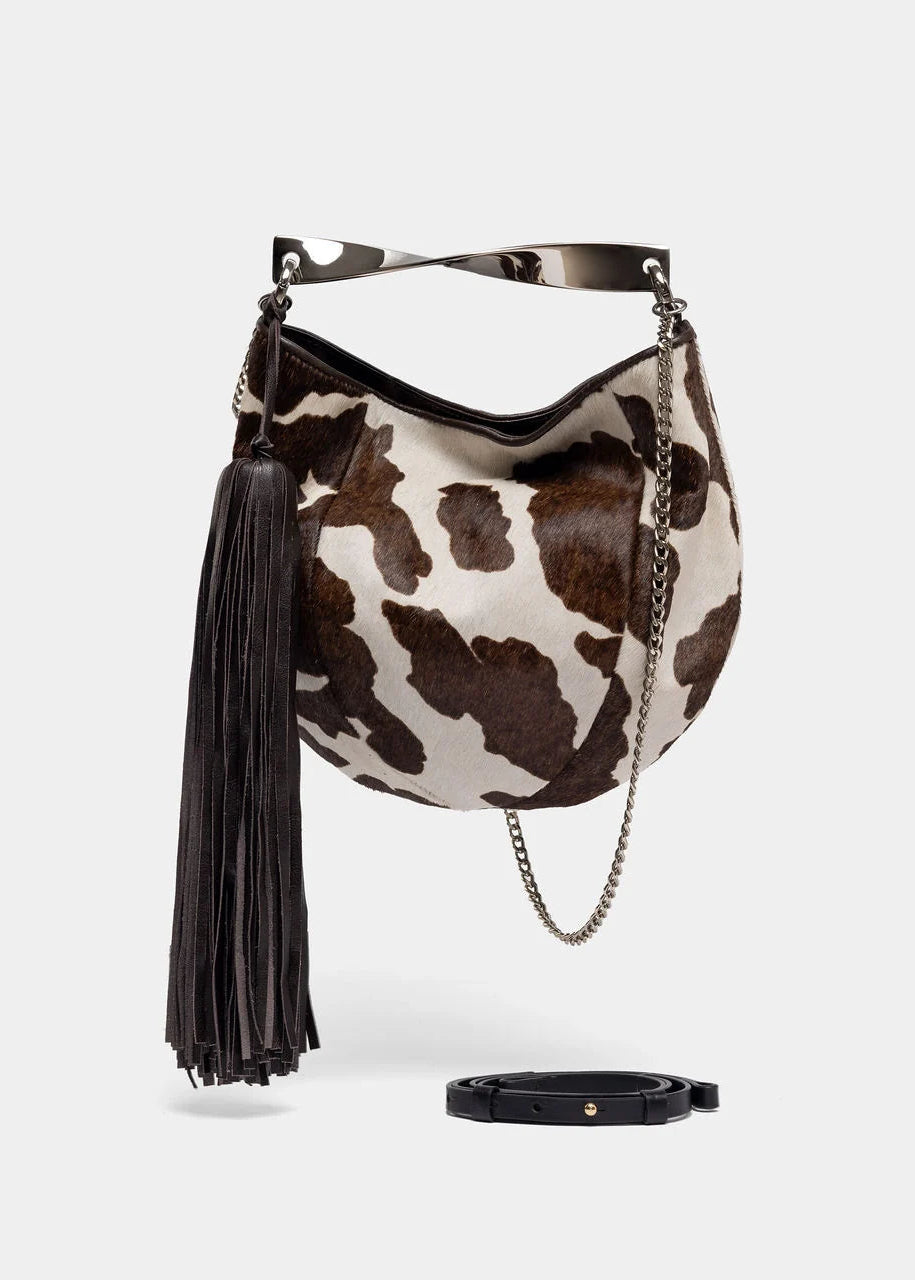 A crescent-shaped shoulder bag with a brown and white cow print, featuring a black leather tassel, a silver-tone metal handle, and a silver chain strap. A black leather belt is shown below the bag on a light gray background.