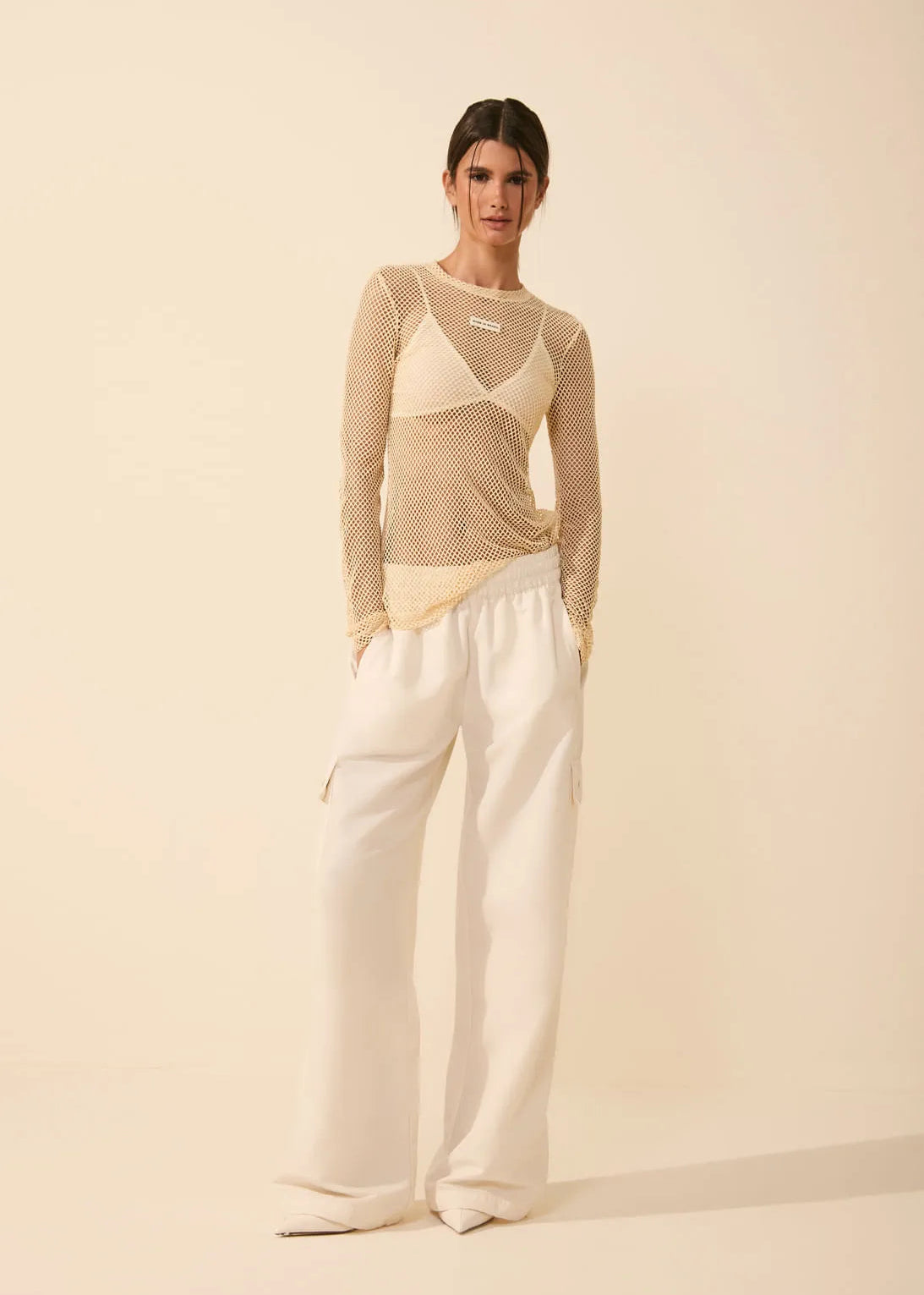 Woman wearing a beige long-sleeve top and white pants against a beige background