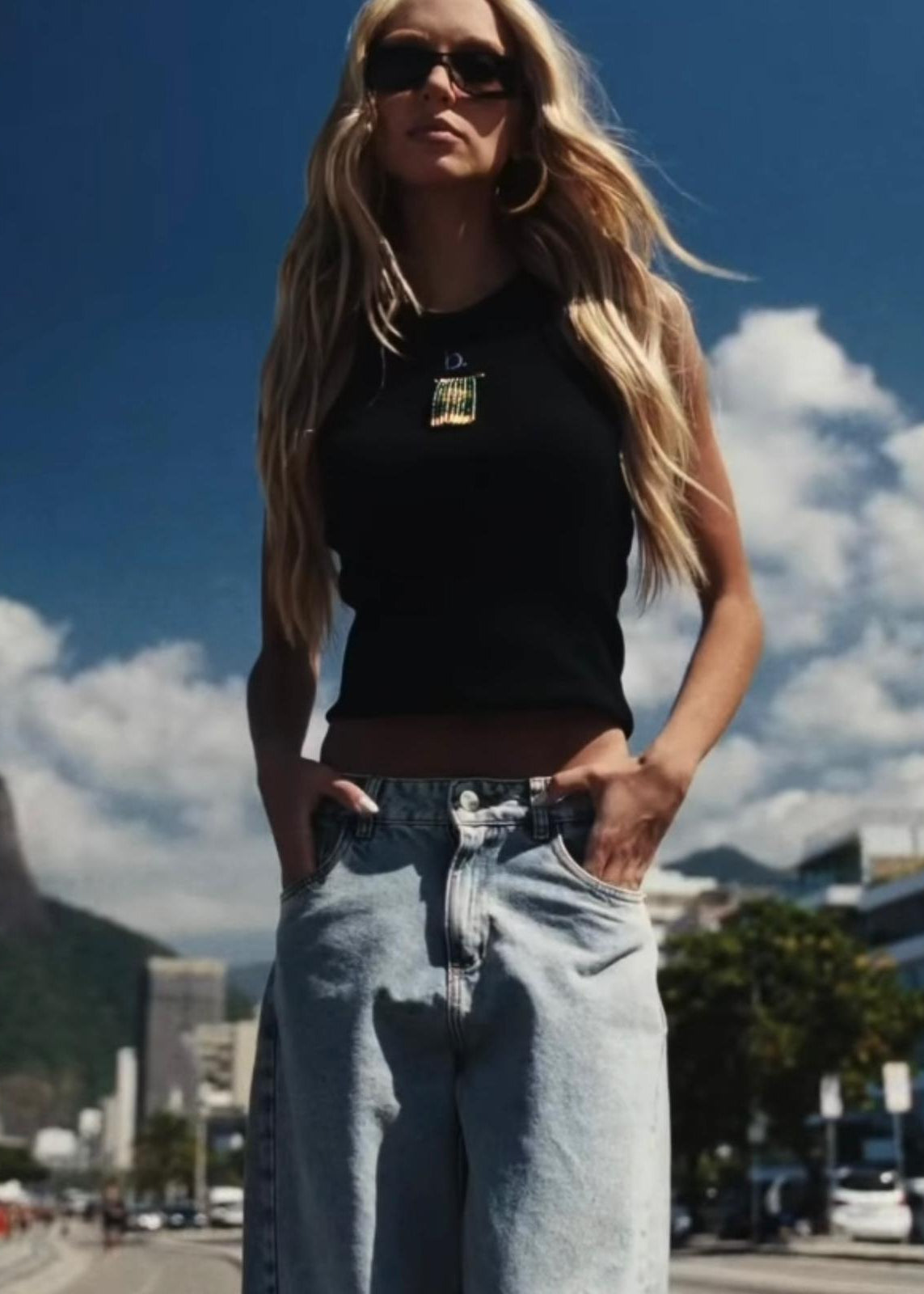 Woman wearing a black top and light blue jeans with sunglasses, standing outdoors against a cityscape.