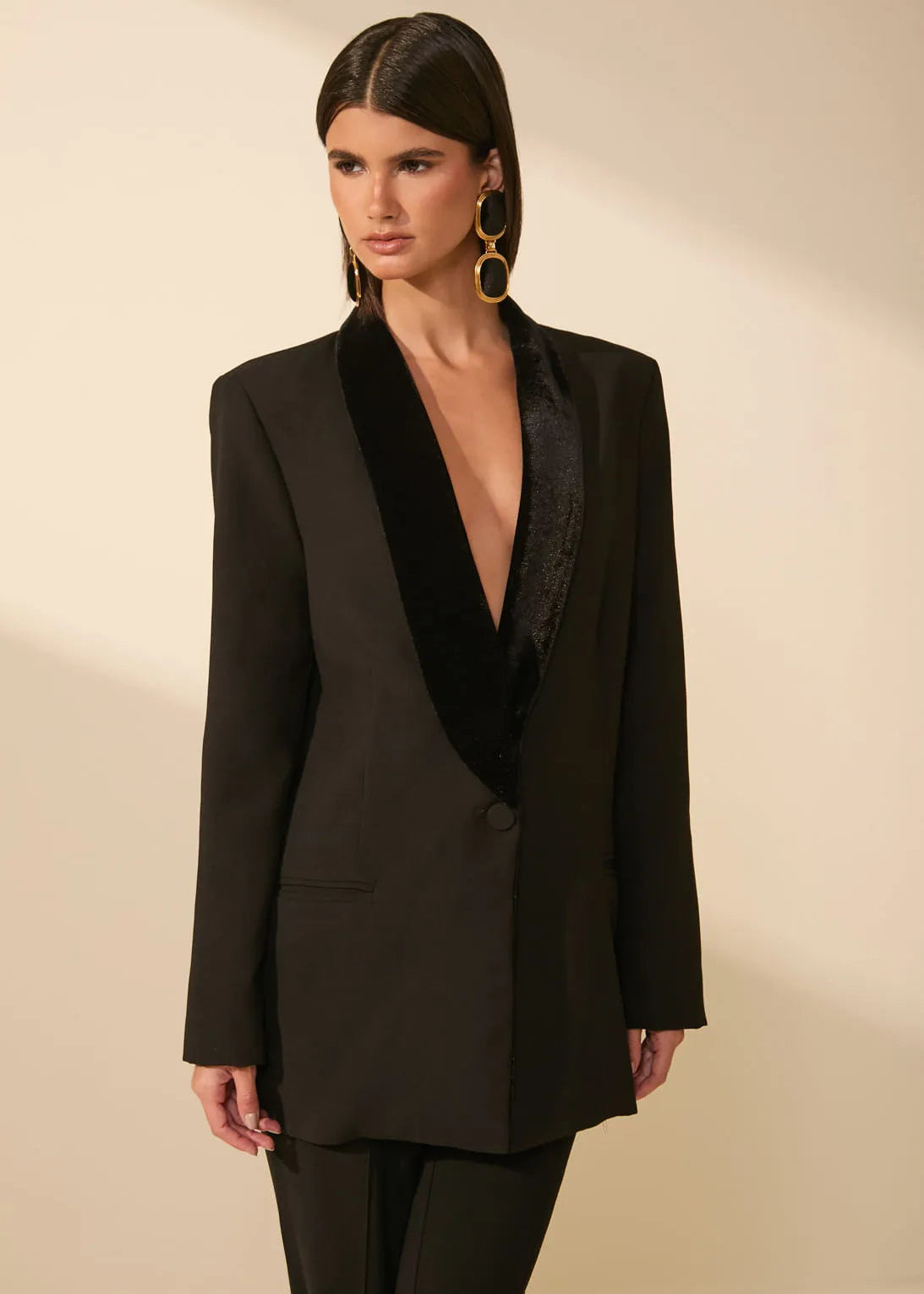 A model wearing a black blazer with a contrasting black velvet shawl collar, paired with matching black trousers and gold hoop earrings, set against a warm beige studio background.