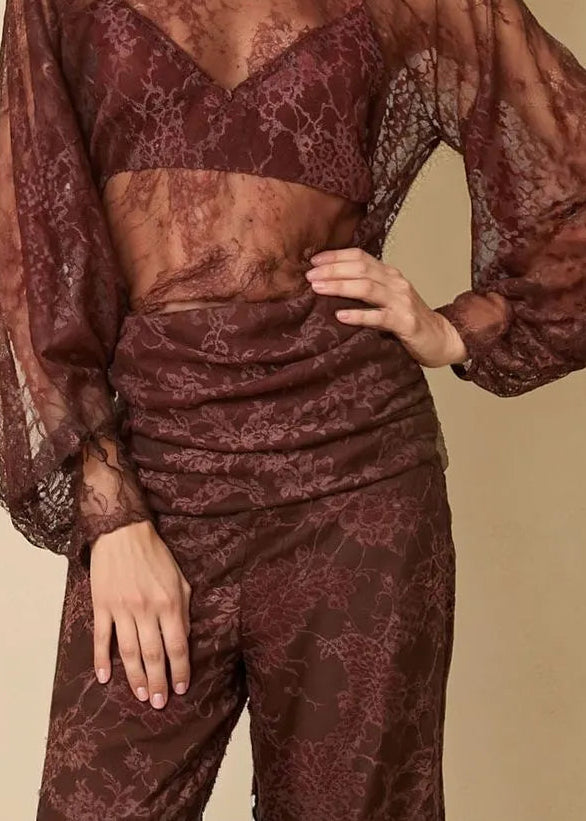Woman wearing a sheer maroon top and pants with a beige hat on a beige background