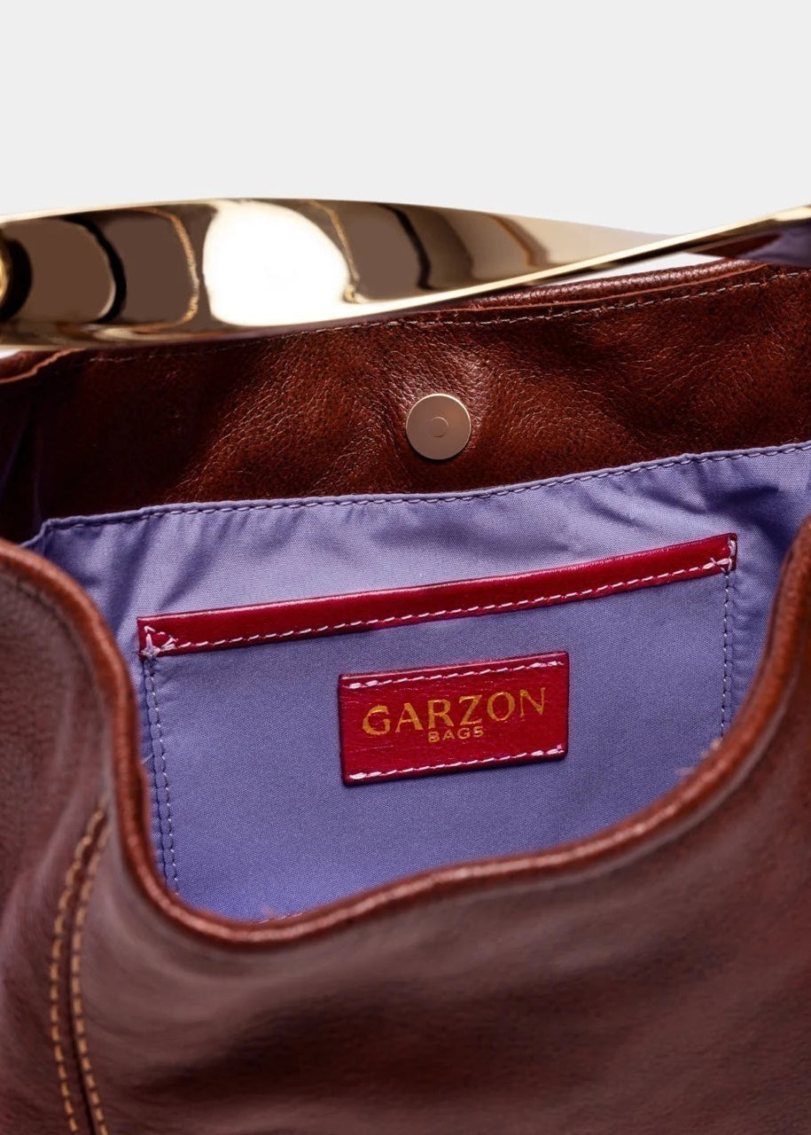 Close-up of a brown leather handbag interior showing a lavender blue satin lining with a red pocket and a GARZON MADE brand label.