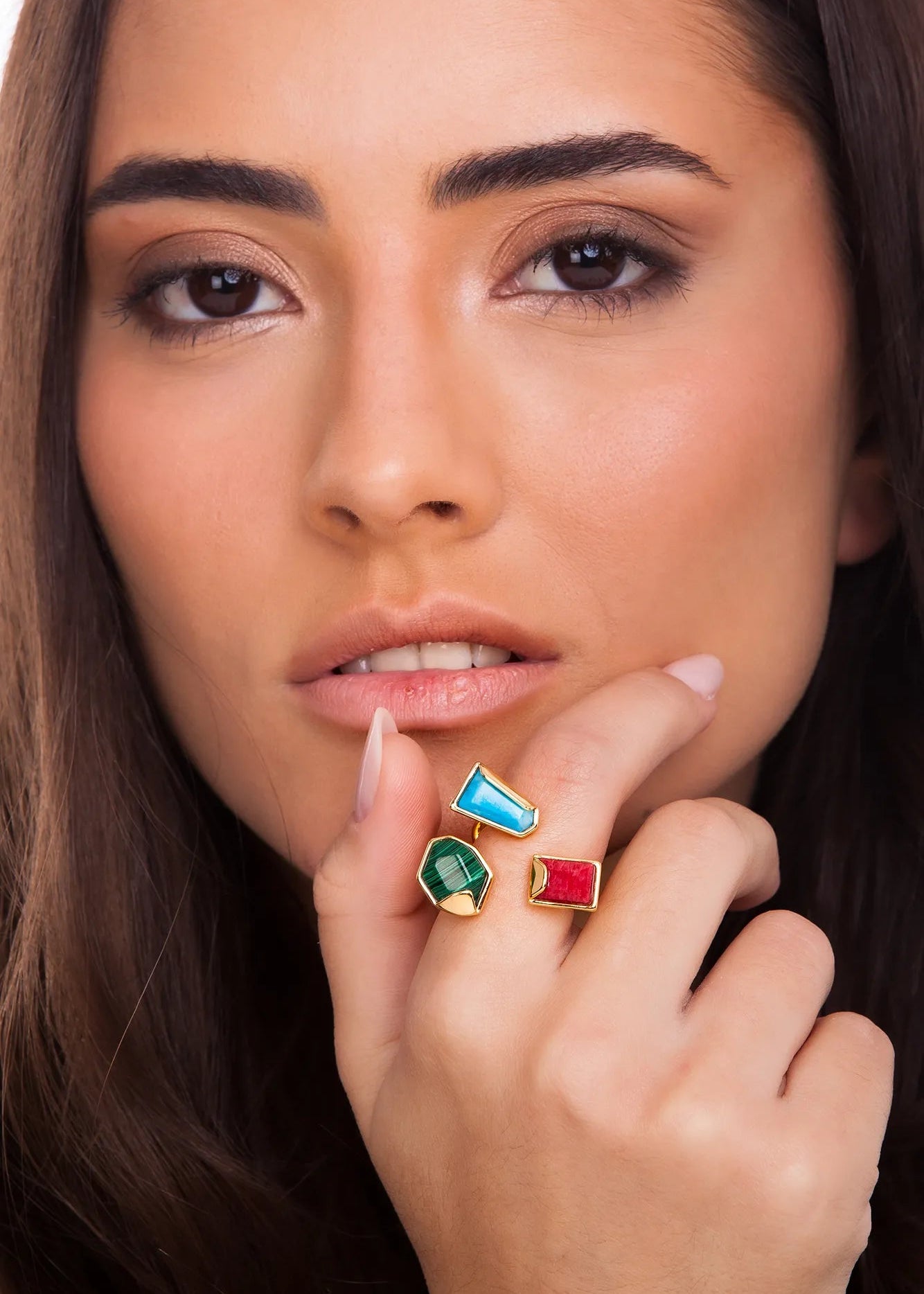Close-up of a woman's face with colorful gemstone rings on her fingers.