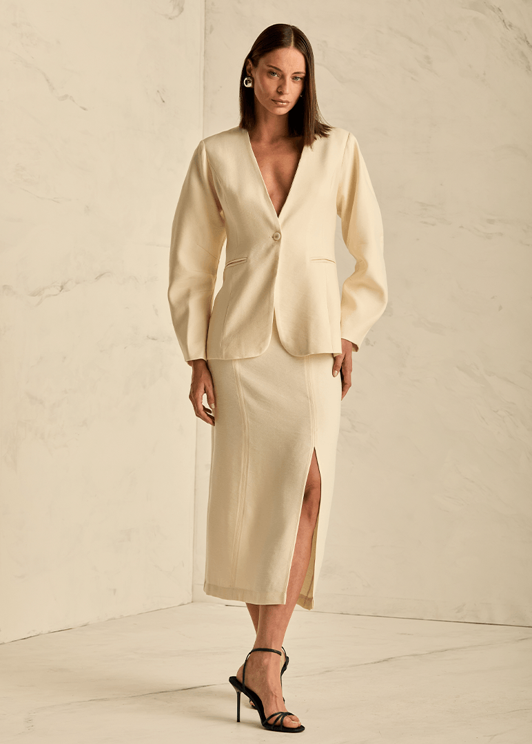 Woman wearing a beige suit with a deep neckline and high slit skirt against a neutral background