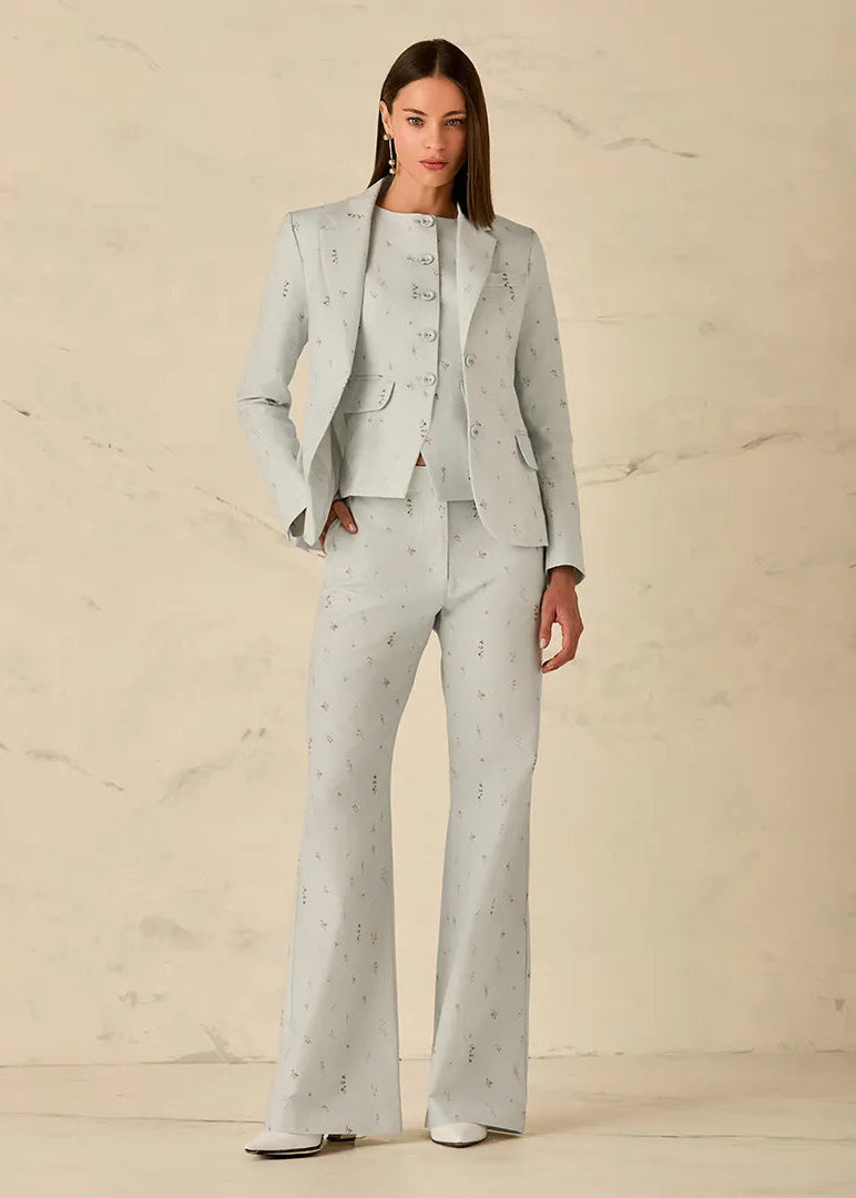 A model stands against a light beige marble-textured wall wearing a light gray three-piece suit with a subtle black floral pattern, consisting of a blazer, vest, and wide-leg trousers.