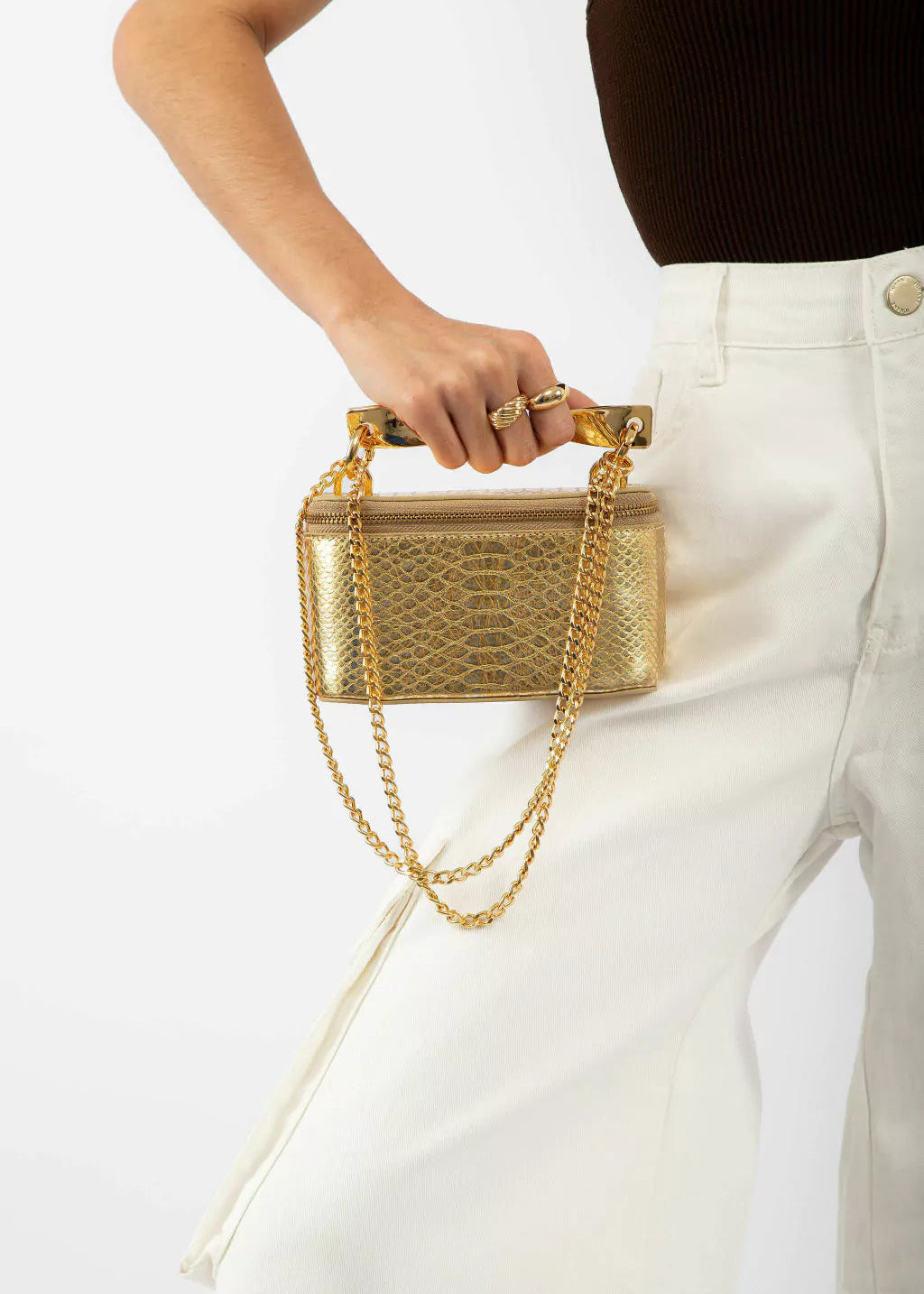 Gold handbag with chain detail held by a person wearing white pants on a light background