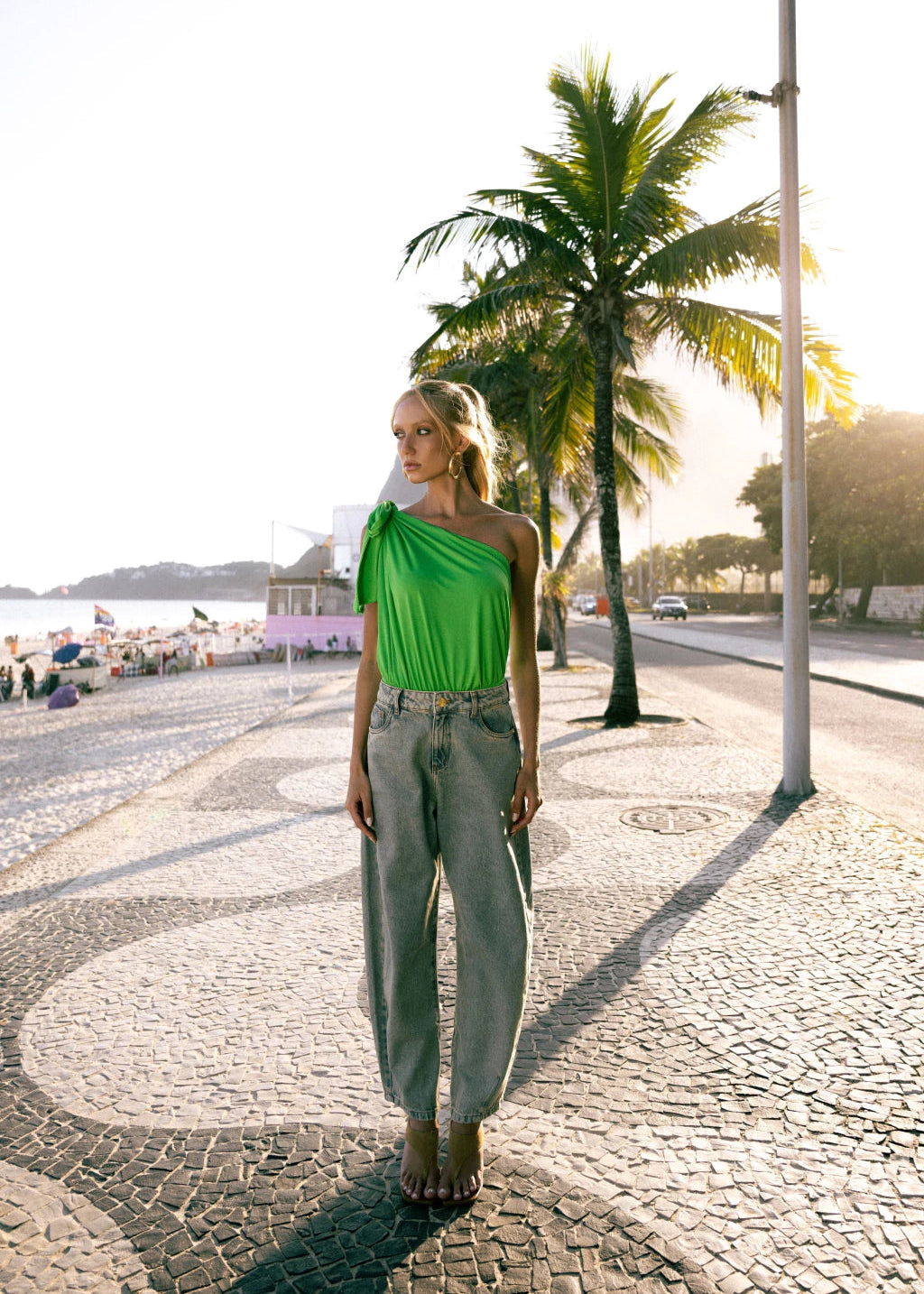 Woman in a green top and gray pants standing on a sunlit street with palm trees.