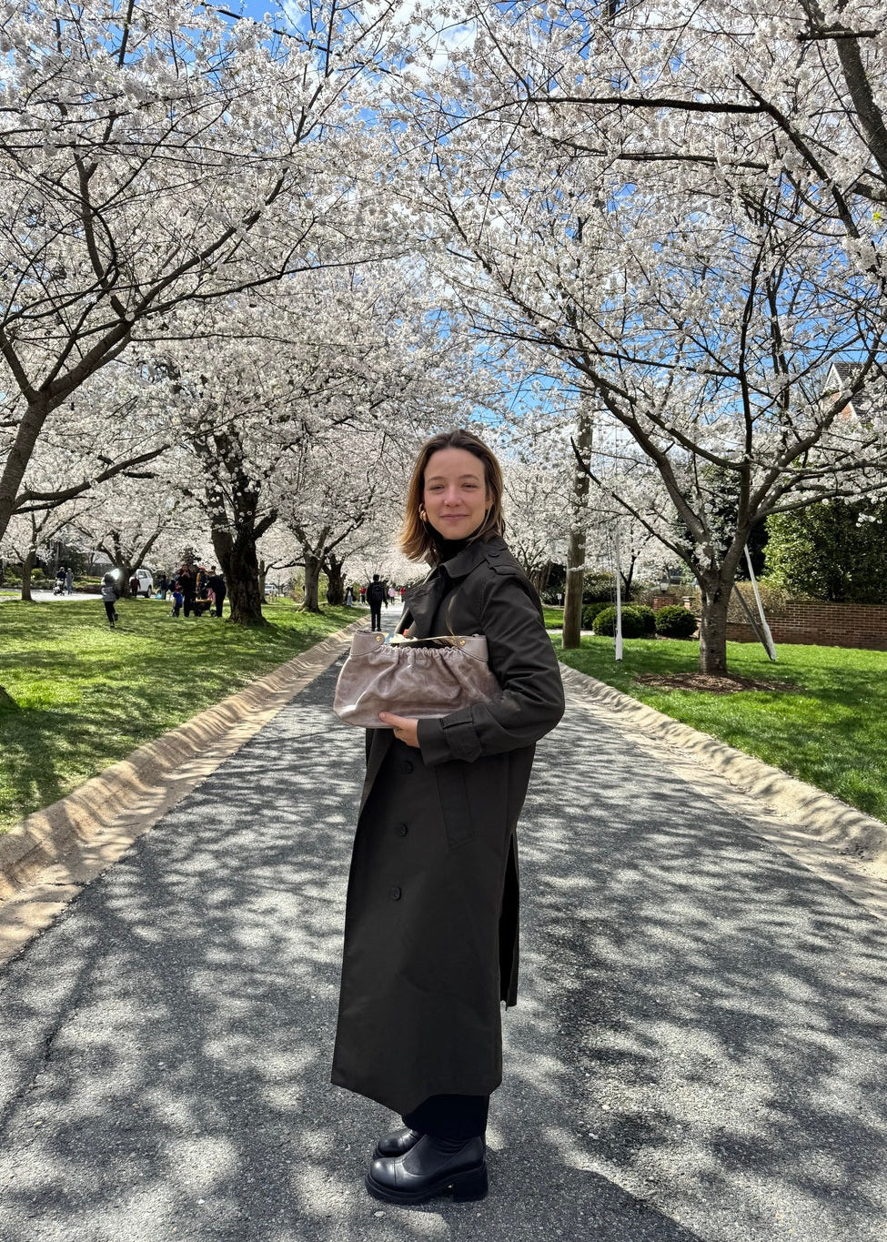 A woman stands on a paved path surrounded by blooming cherry blossom trees, wearing a long dark coat and holding a light-colored handbag.