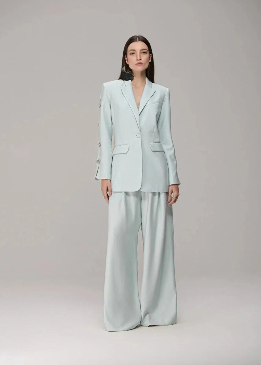 A model stands against a neutral light gray studio background wearing a light mint green two-piece suit consisting of a single-breasted blazer with wide lapels and matching wide-leg trousers.