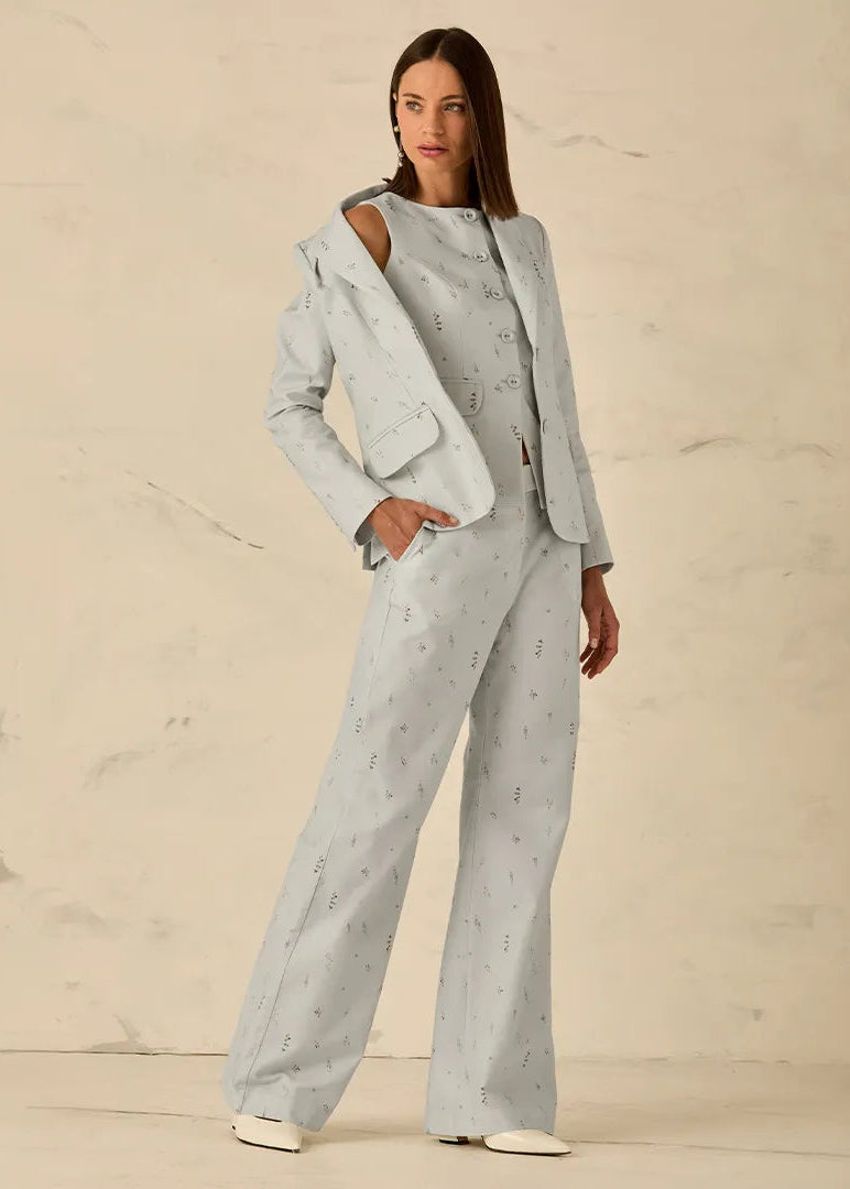 A woman models a light gray two-piece suit with a subtle geometric pattern, featuring a collared jacket with shoulder cutouts and matching wide-leg trousers, set against a textured beige studio backdrop.