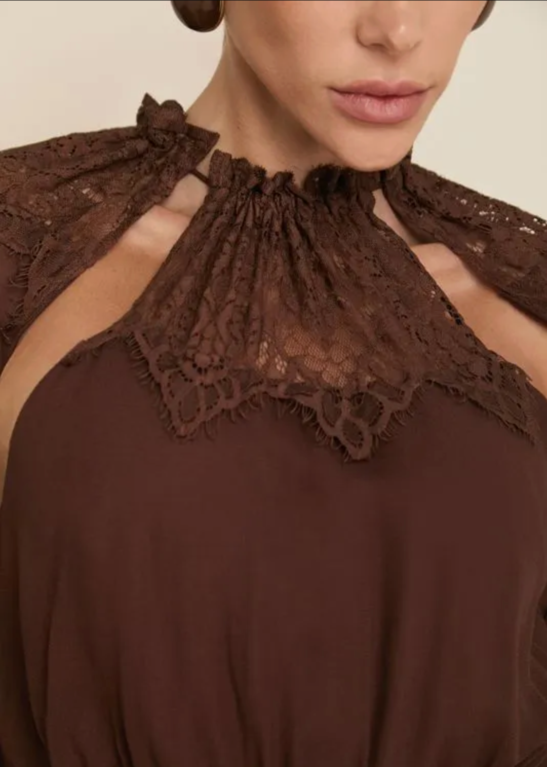 Close-up of a model wearing a dark brown top with a lace high neckline and matching lace shoulder ties against a light beige background.