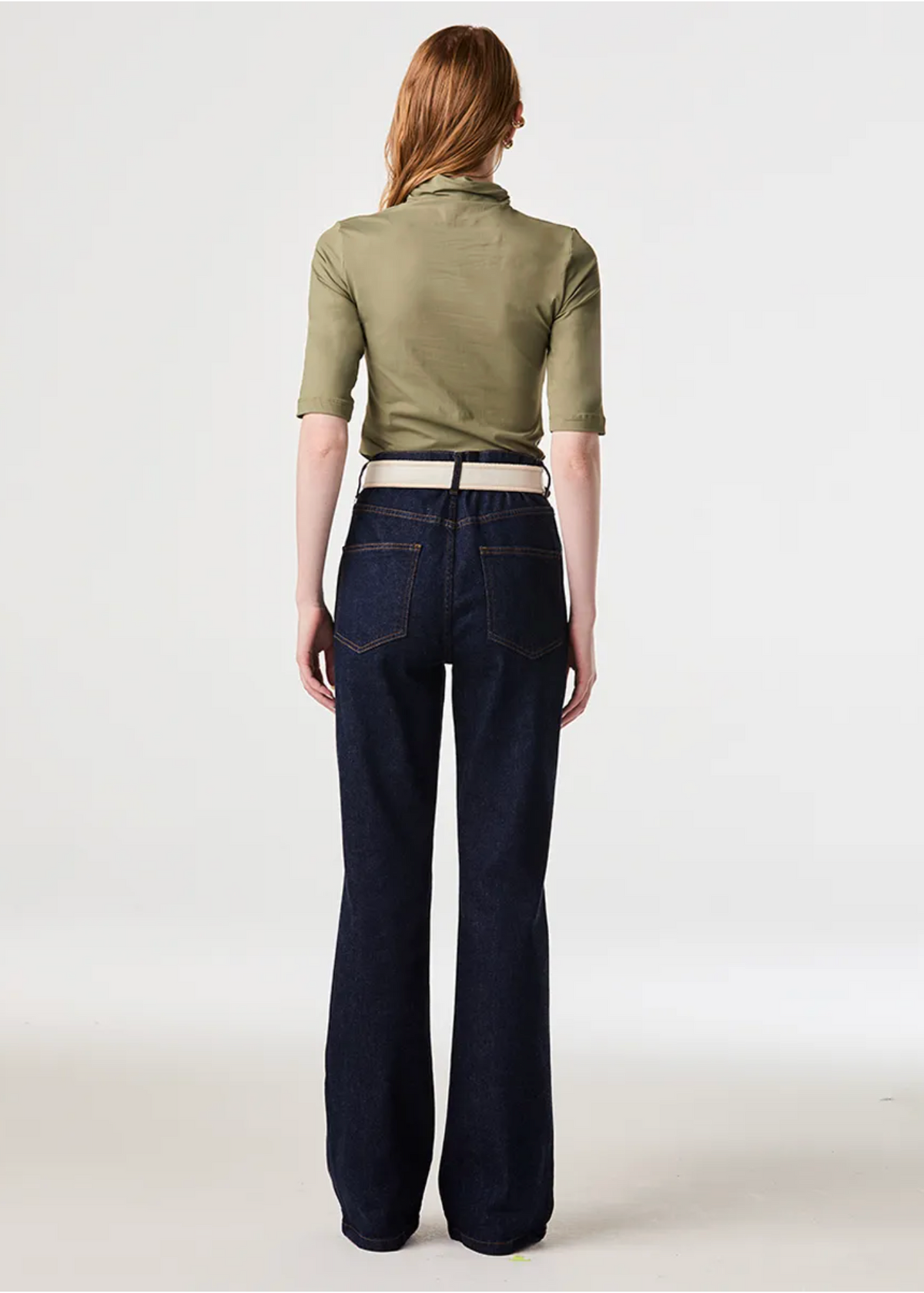 A woman viewed from the back wearing an olive green short-sleeve mock-neck top and dark blue wide-leg jeans with a light beige belt, set against a plain light gray studio background.