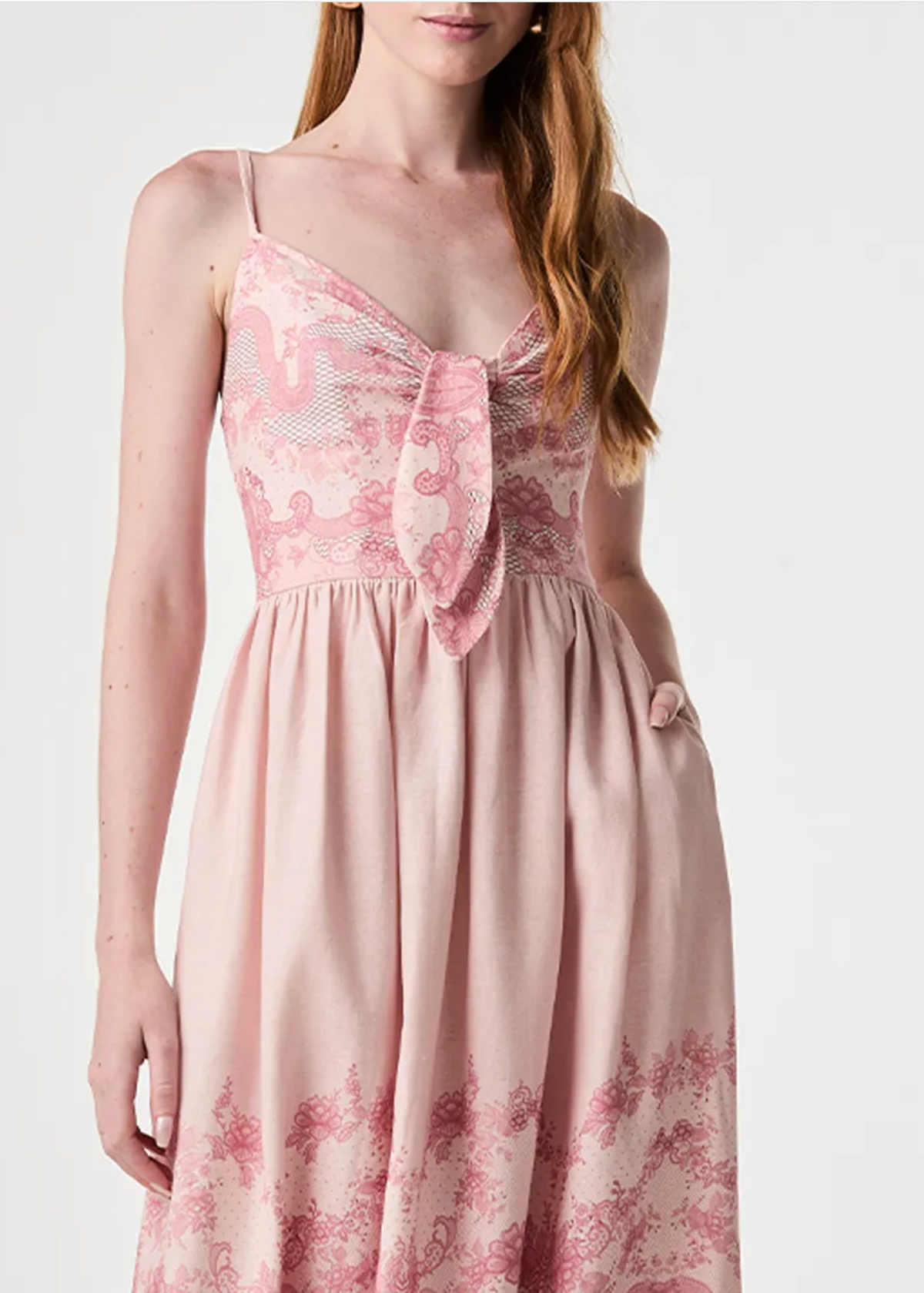A woman wearing a light pink spaghetti-strap dress with a tie-front bodice and a pink floral toile pattern, shown against a plain light gray studio background.