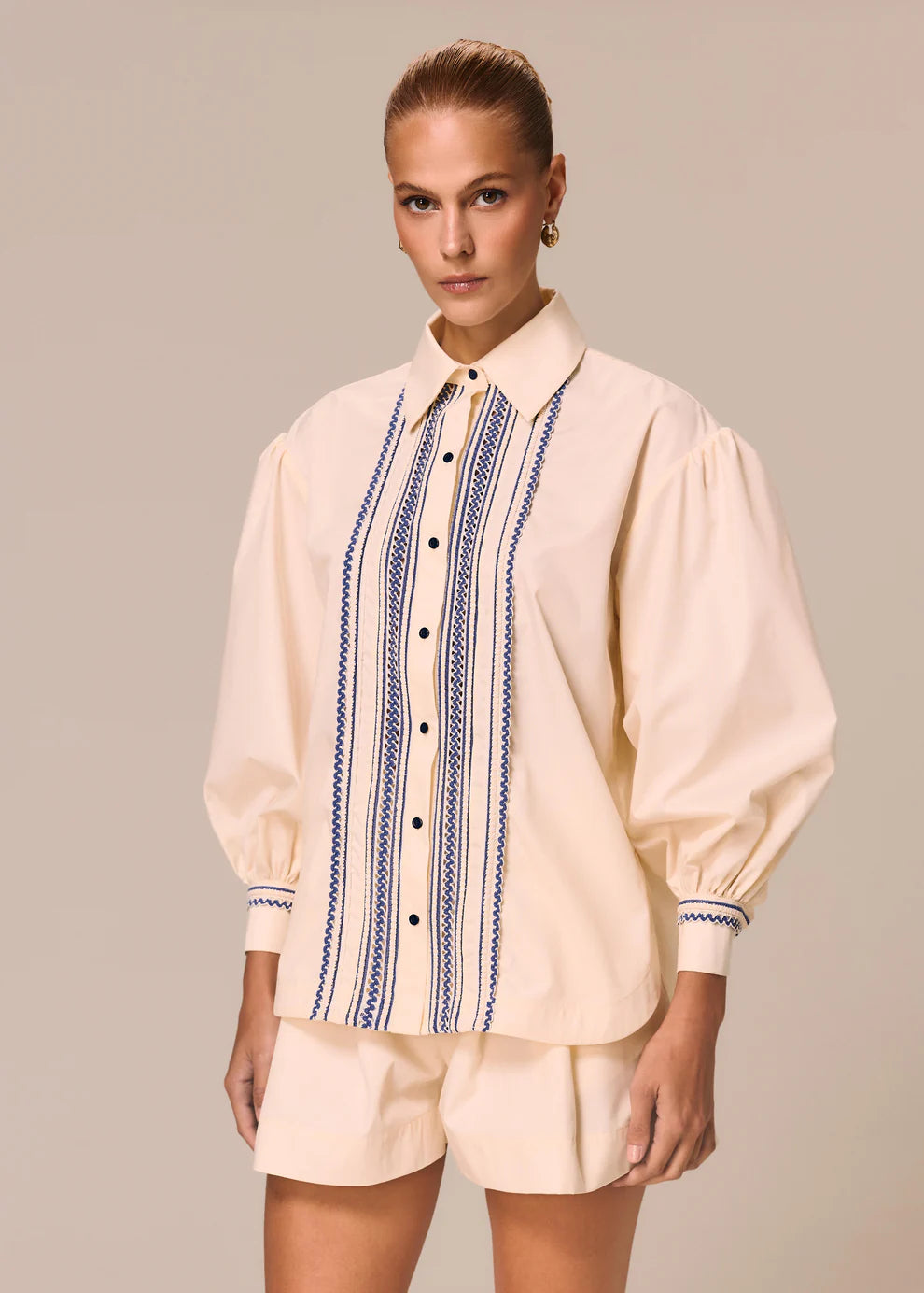 Woman wearing a cream blouse with blue embroidery and matching shorts on a beige background