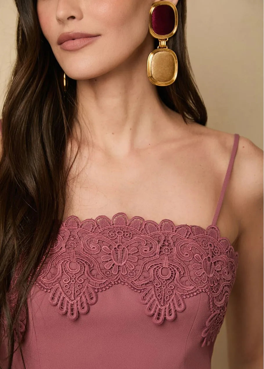 Close-up of a woman wearing a pink lace top with gold earrings against a beige background