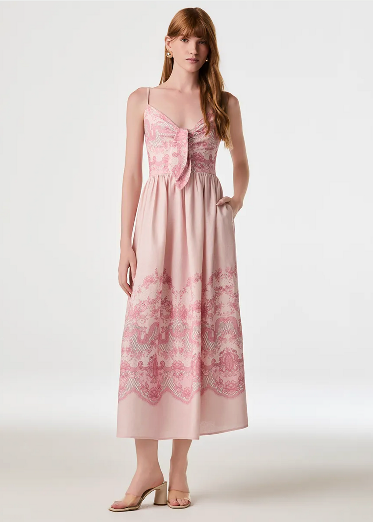 A woman modeling a light pink midi dress with floral lace detailing on the bodice and hem, photographed against a plain light gray studio background.