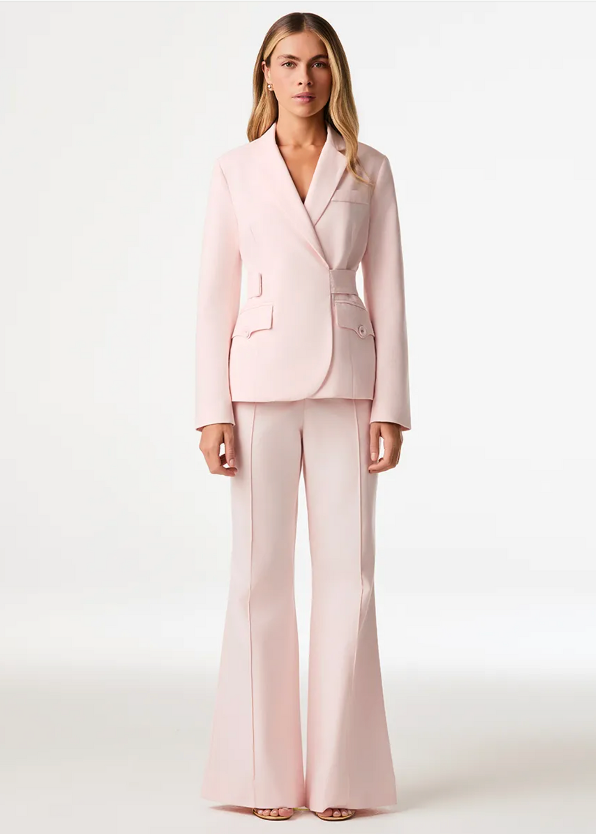 A woman stands against a clean white studio background wearing a light pink tailored suit with a double-breasted blazer and matching wide-leg pants.