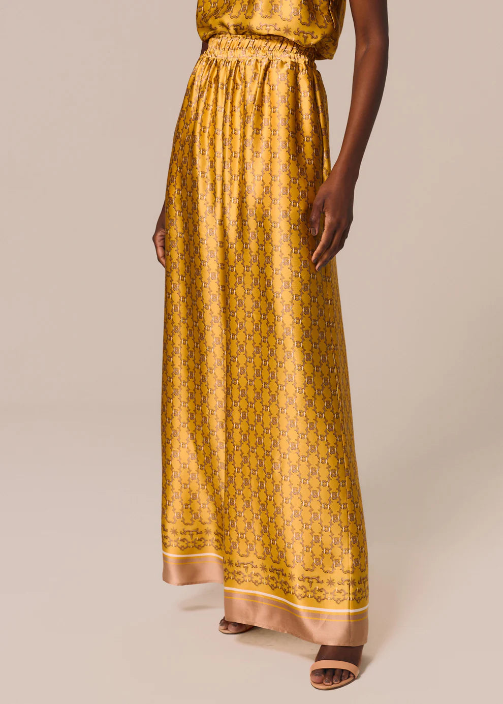 A model wearing a long, wide-leg yellow maxi skirt with a repeating geometric pattern and a solid border, paired with brown heeled sandals against a solid beige background.